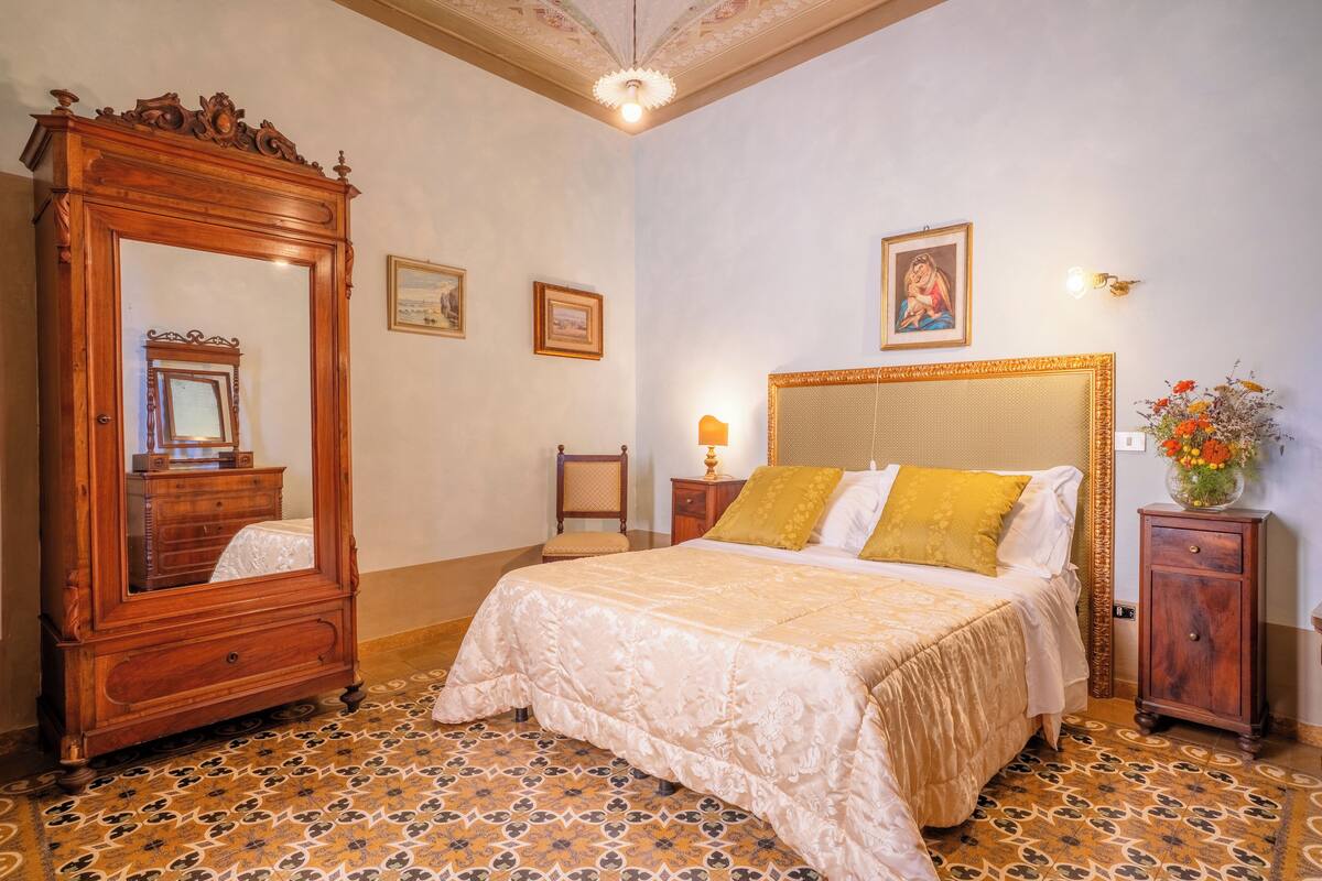 Une chambre à coucher avec un grand miroir décoratif, un lit agrémenté d’un coussin jaune, une table de chevet avec une lampe et une petite armoire.