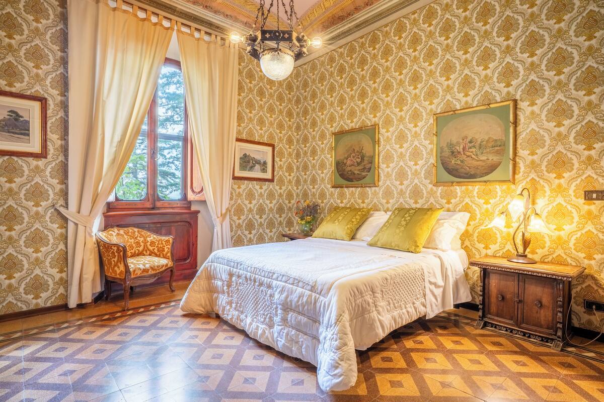 Une chambre avec un papier peint à motifs, un grand lit recouvert d’une courtepointe blanche, une table de chevet en bois avec une lampe et une chaise.