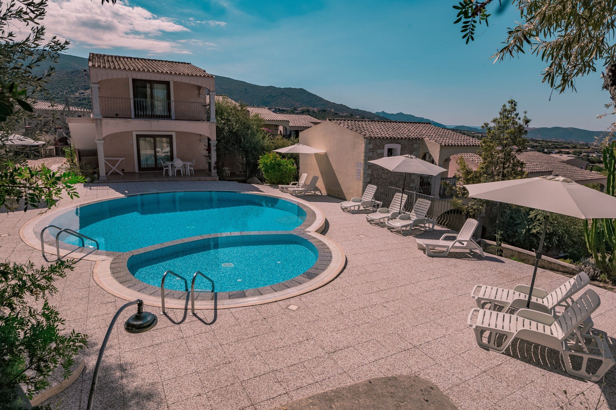 A swimming pool with lounge chairs and umbrellas, surrounded by buildings with a mountainous backdrop.