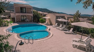 A swimming pool with lounge chairs and umbrellas, surrounded by buildings with a mountainous backdrop.
