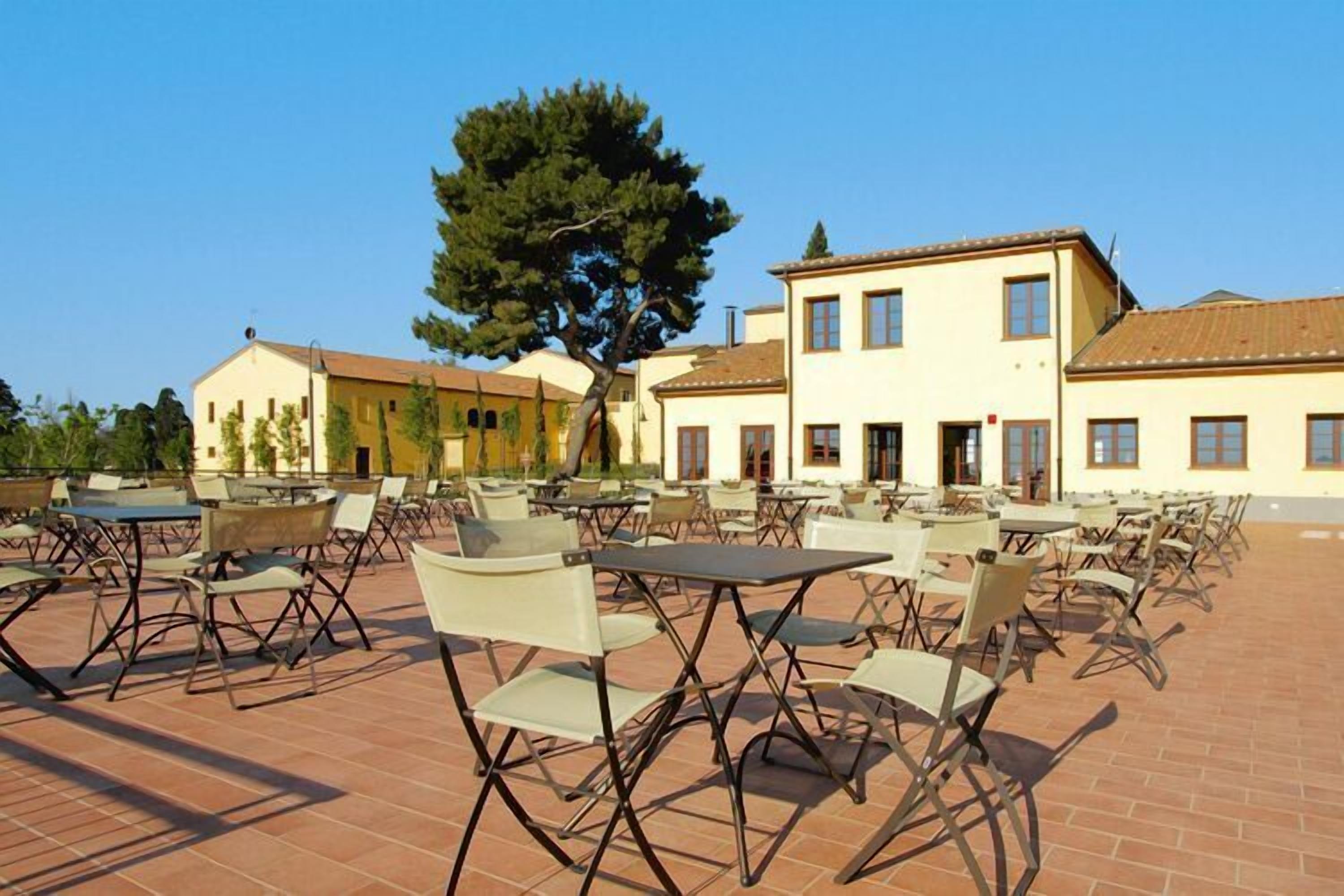 An outdoor seating area with tables and chairs, a large tree, and buildings in the background.