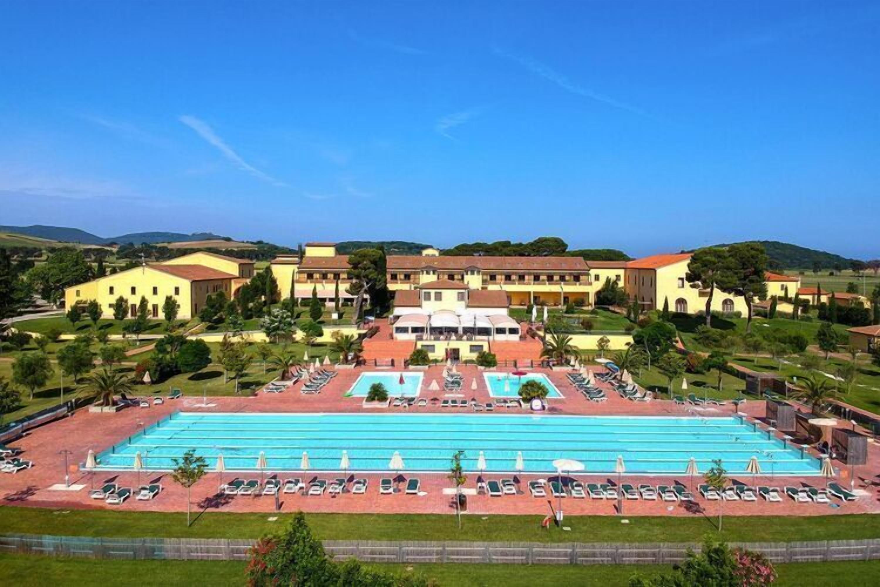 A large swimming pool with multiple lanes, surrounded by lounge chairs and umbrellas, with a building complex in the background.