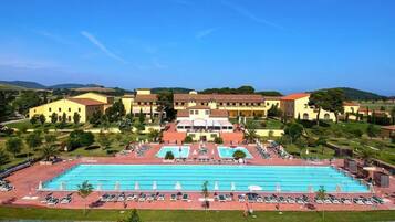 A large swimming pool with multiple lanes, surrounded by lounge chairs and umbrellas, with a building complex in the background.