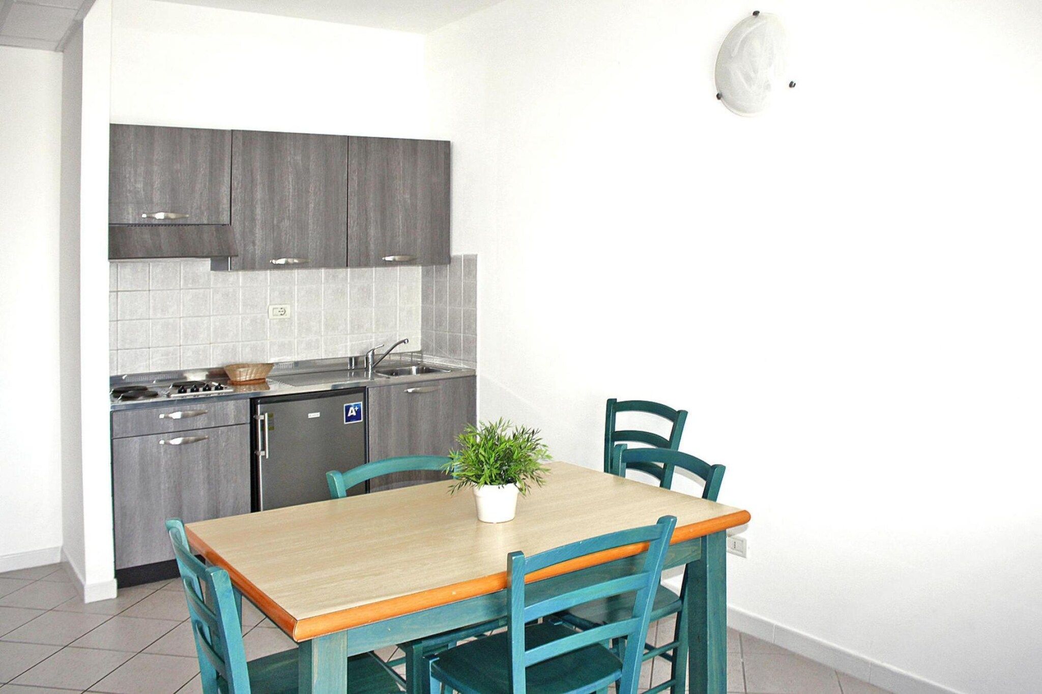 A compact kitchen with grey cabinets, a stainless steel stove, and a small sink. There is a table with two chairs and a potted plant on it.