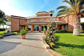 A hotel with a brick facade, a covered entrance, and a well-maintained garden with a palm tree and flowering plants. - Villa in Cecina Mare near Sandy Beach (Cecina Mare)