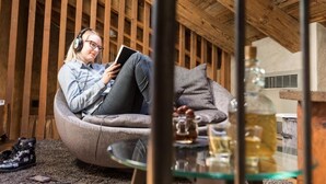 Iemand zit in een krukkonenbank, een boek lezend in een kamer met houten balken aan het plafond en een glazen tafel met flessen.