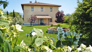 Une maison à deux étages, avec un toit en tuiles, un petit porche et un jardin bien entretenu où poussent des plantes à fleurs.