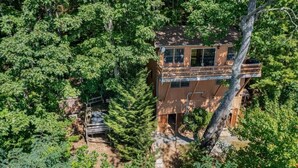 A treehouse with a wooden exterior, a balcony, and a staircase leading up to it, surrounded by dense forest. - Skyview Cabin - Spectacular View, 2 BR, 2 1/2 BA, Brand New Hot Tub, Firepit (Maggie Valley)