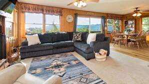 A cozy living room with a sectional sofa, a dining table with chairs, a ceiling fan, and a large window with a view of the outdoors. - Skyview Cabin - Spectacular View, 2 BR, 2 1/2 BA, Brand New Hot Tub, Firepit (Maggie Valley)