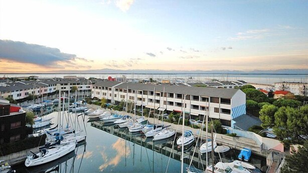 A marina with multiple boats docked, a building with a checkered roof, and a view of the sea and distant land.