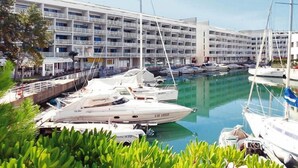 A marina with multiple yachts docked, a large hotel building with balconies, and lush greenery in the foreground.