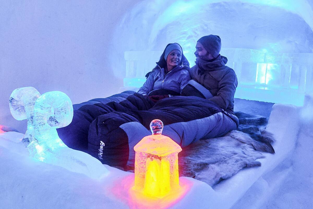 A couple sitting on a bed in an igloo with ice sculptures and a lantern.