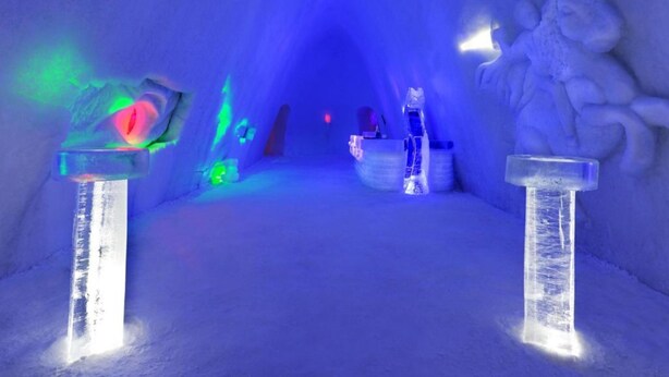 An ice tunnel with a person standing in the center, illuminated by blue and green lights, and ice sculptures on either side. - Hunderfossen Snøhotell (Lillehammer)