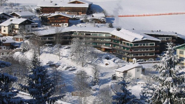A snow-covered resort with multiple buildings, including a large hotel with a curved facade and a smaller building with a sign. - Apartment in Wildschönau by Schatzberg Lift (Wildschönau)