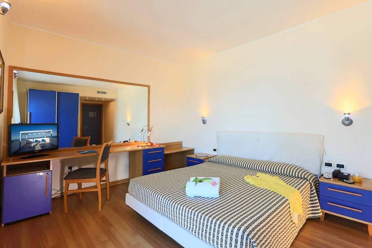 A hotel room with a bed, a desk with a computer, a chair, a small fridge, and a mirror.