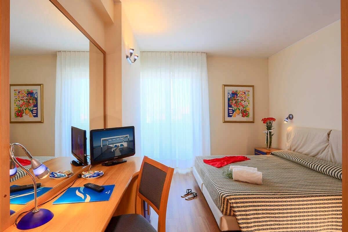 A hotel room with a bed, a desk with a computer, a chair, and a mirror.
