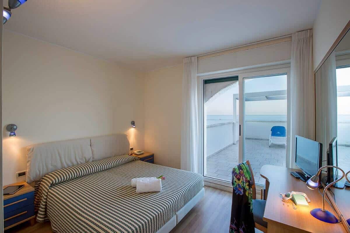 A hotel room with a bed, a desk with a computer, a chair, and a large window with a view of the beach.