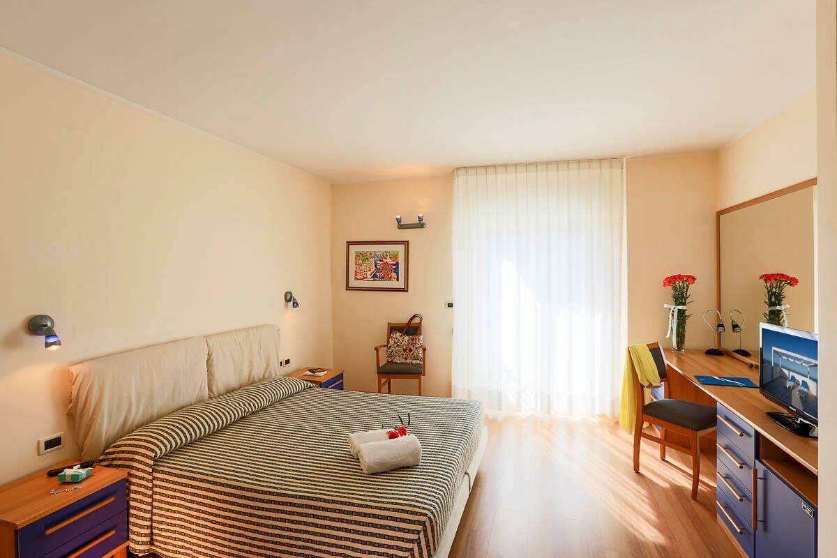 A hotel room with a large bed, a desk, a chair, a TV, a desk lamp, a bedside table, and a window with curtains.
