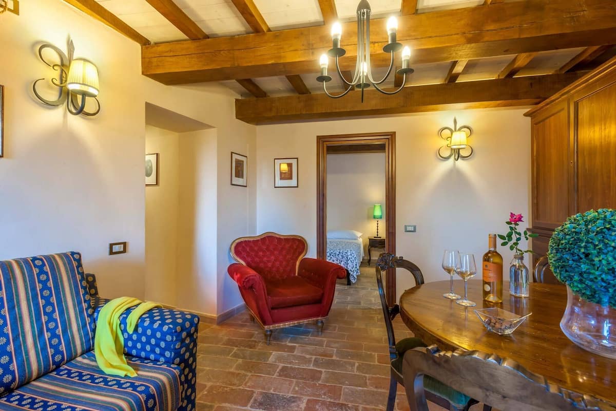 Una habitación acogedora con techo de madera, un sillón rojo, un sofá estampado y una mesa de comedor con copas de vino y una botella.