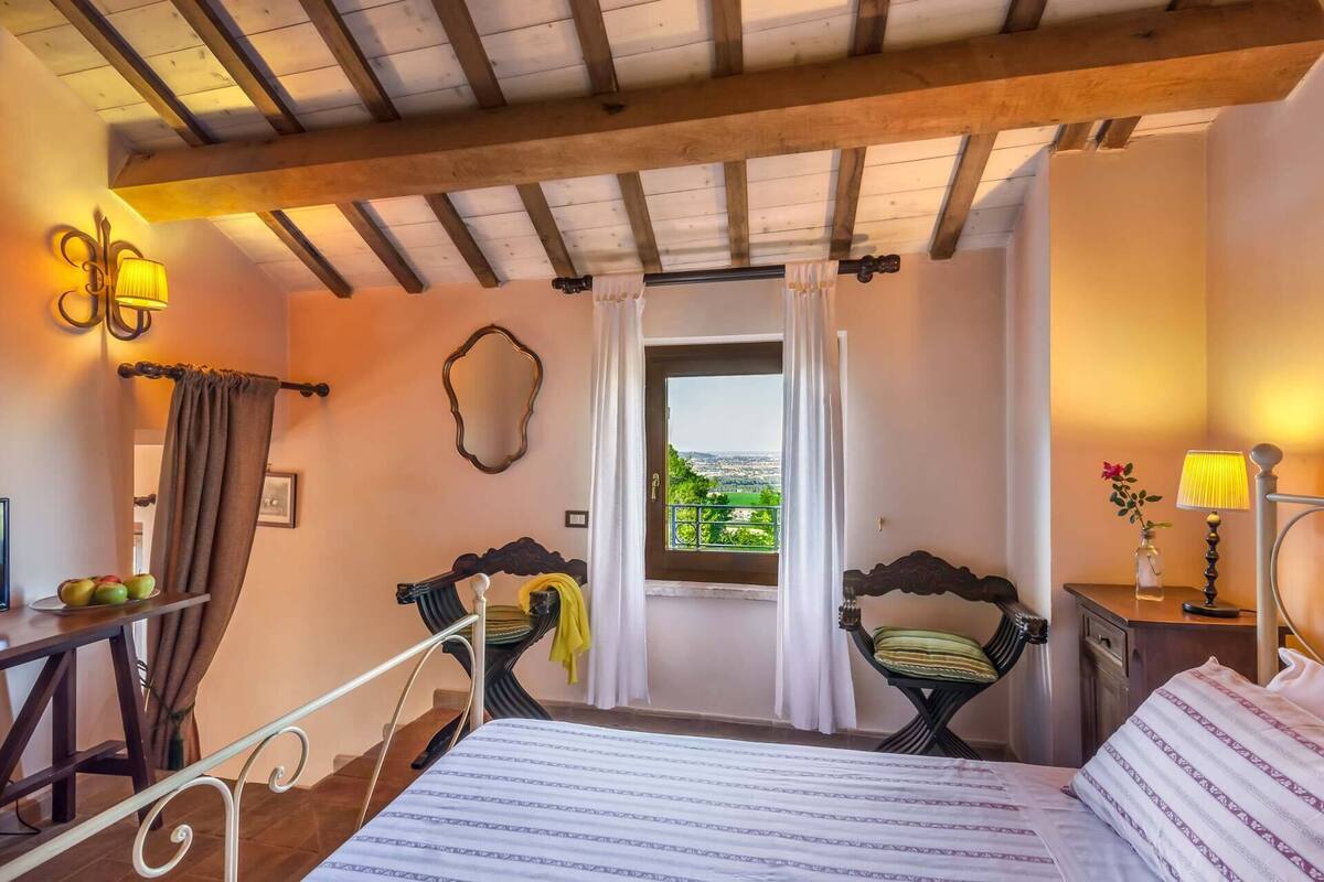 Un dormitorio con cama, dos sillas, una mesa con fruta, ventana con vistas y una lámpara.