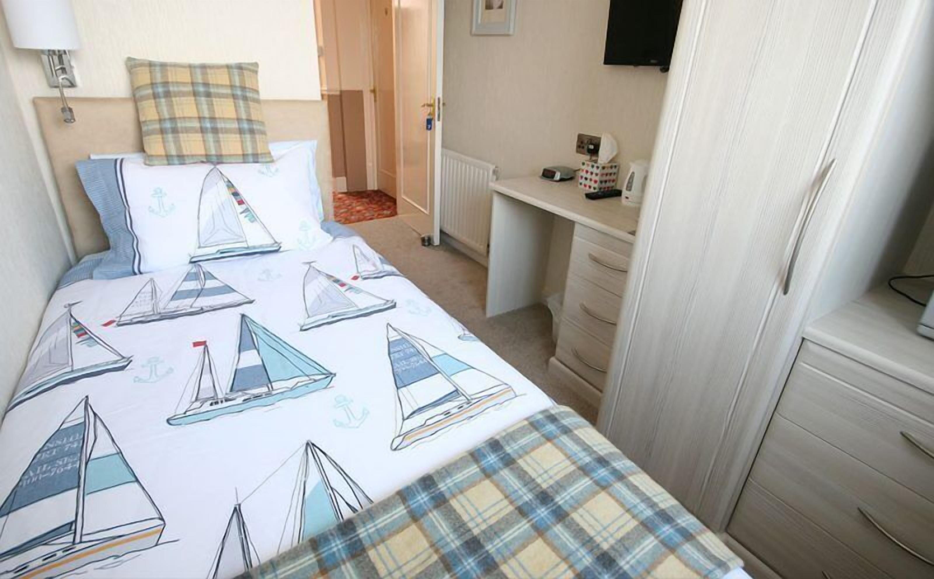 A bedroom with a bed featuring sailboat designs, a headboard, a bedside table with a lamp, a dresser, and a desk with a chair.