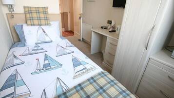 A bedroom with a bed featuring sailboat designs, a headboard, a bedside table with a lamp, a dresser, and a desk with a chair.