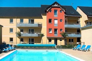 Une piscine avec des chaises longues, un bâtiment doté de balcons et un ciel d’un bleu limpide.