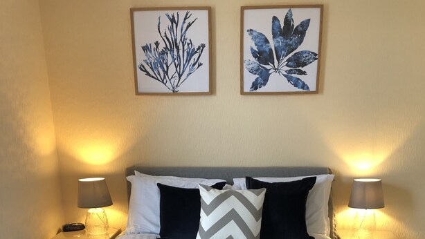 A neatly made bed with a blue bedspread, two white towels, and decorative pillows. There are two bedside lamps and framed artwork on the wall. - Mounts Bay Inn (Helston)