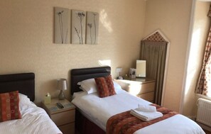 A hotel room with two beds, a nightstand with a lamp, a mirror, and a window with curtains. - Mounts Bay Inn (Helston)