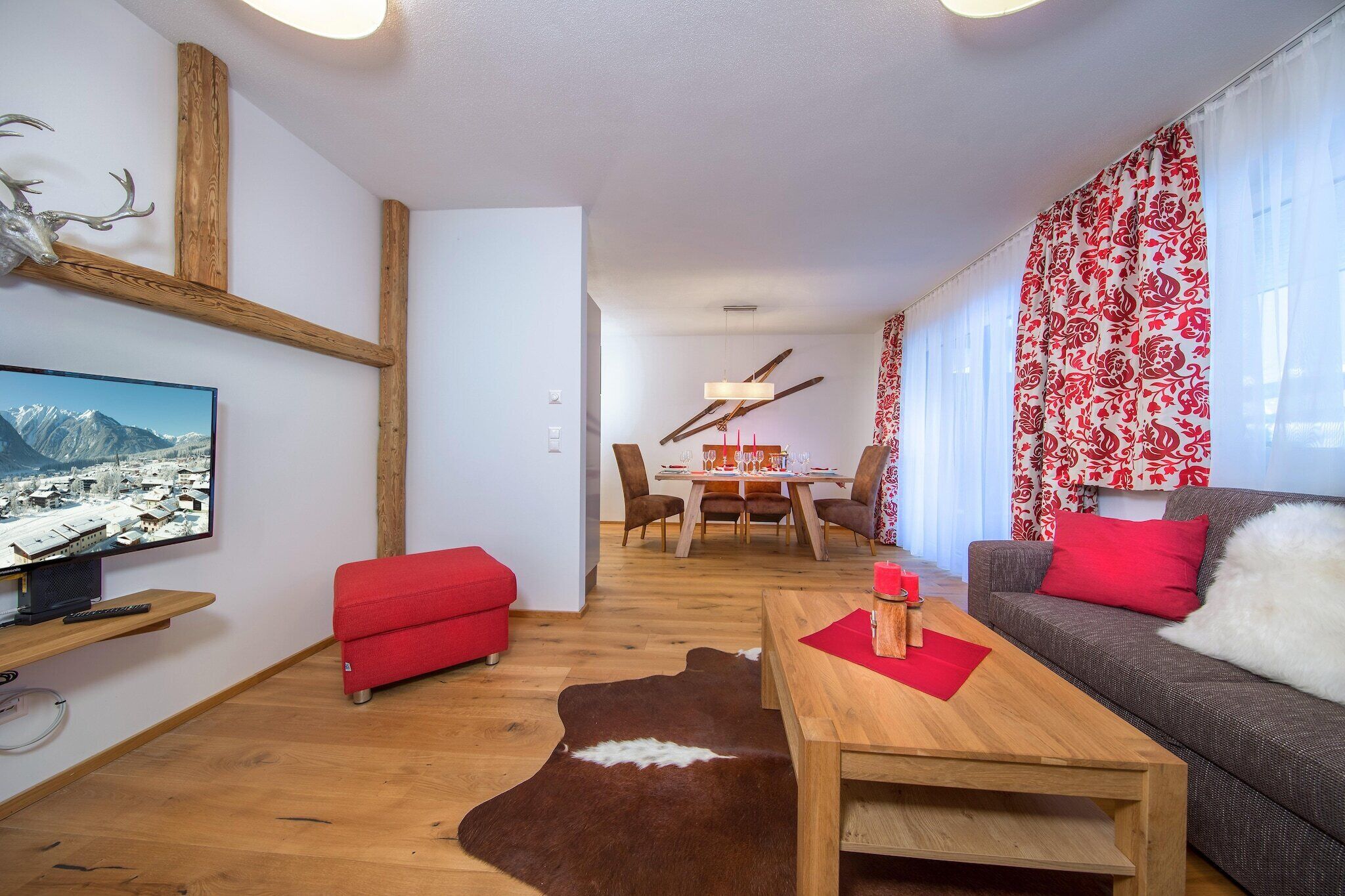 Ein Wohnzimmer mit Couchtisch aus Holz, einem roten Sofa und einem roten Ottomanen. Ein Fernseher steht auf einem Holzregal, ein Esstisch mit Stühlen und ein Kuhfellteppich sind ebenfalls zu sehen.