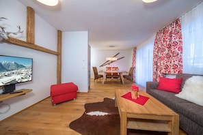 A living room with a wooden coffee table, red sofa, and a red ottoman. There is a television on a wooden shelf, a wooden dining table with chairs, and a cowhide rug.