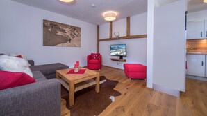 A modern living room with a wooden floor, grey sofa, red armchair, and a small table with candles. A television is mounted on the wall, and a painting is visible.