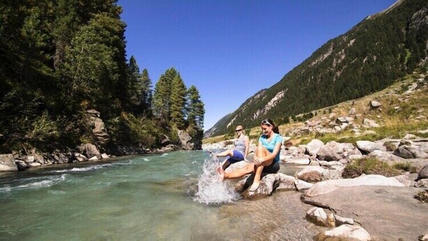 Deux personnes assises sur des rochers, au bord d’une rivière aux eaux claires, avec des montagnes en arrière-plan.