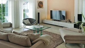 Un salon moderne doté d’un grand téléviseur à écran plat, de canapés beiges, d’une table basse en verre et d’un lustre.