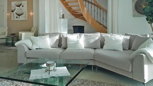 Un salon moderne avec un canapé d’angle beige, une table basse en verre, un escalier en bois et un lustre.