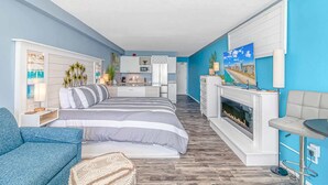A modern bedroom with a bed, a sofa, a fireplace, a TV, and a kitchen area. - Oceanfront King Suite with Sofa~Landmark 830 (Myrtle Beach)