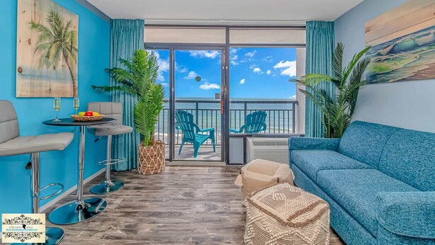 A room with a balcony, a sofa, a table with drinks, and chairs. - Oceanfront King Suite with Sofa~Landmark 830 (Myrtle Beach)