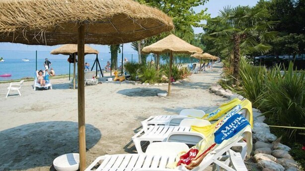 A beach with white lounge chairs and thatched umbrellas, a sandy path, and palm trees. - Mobile home in Cisano San Vito caravan park (Cisano di Bardolino)