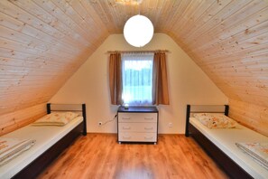 A small, triangular room with wooden walls and a sloped ceiling. It contains two single beds with floral bedding, a small dresser, and a window with curtains.