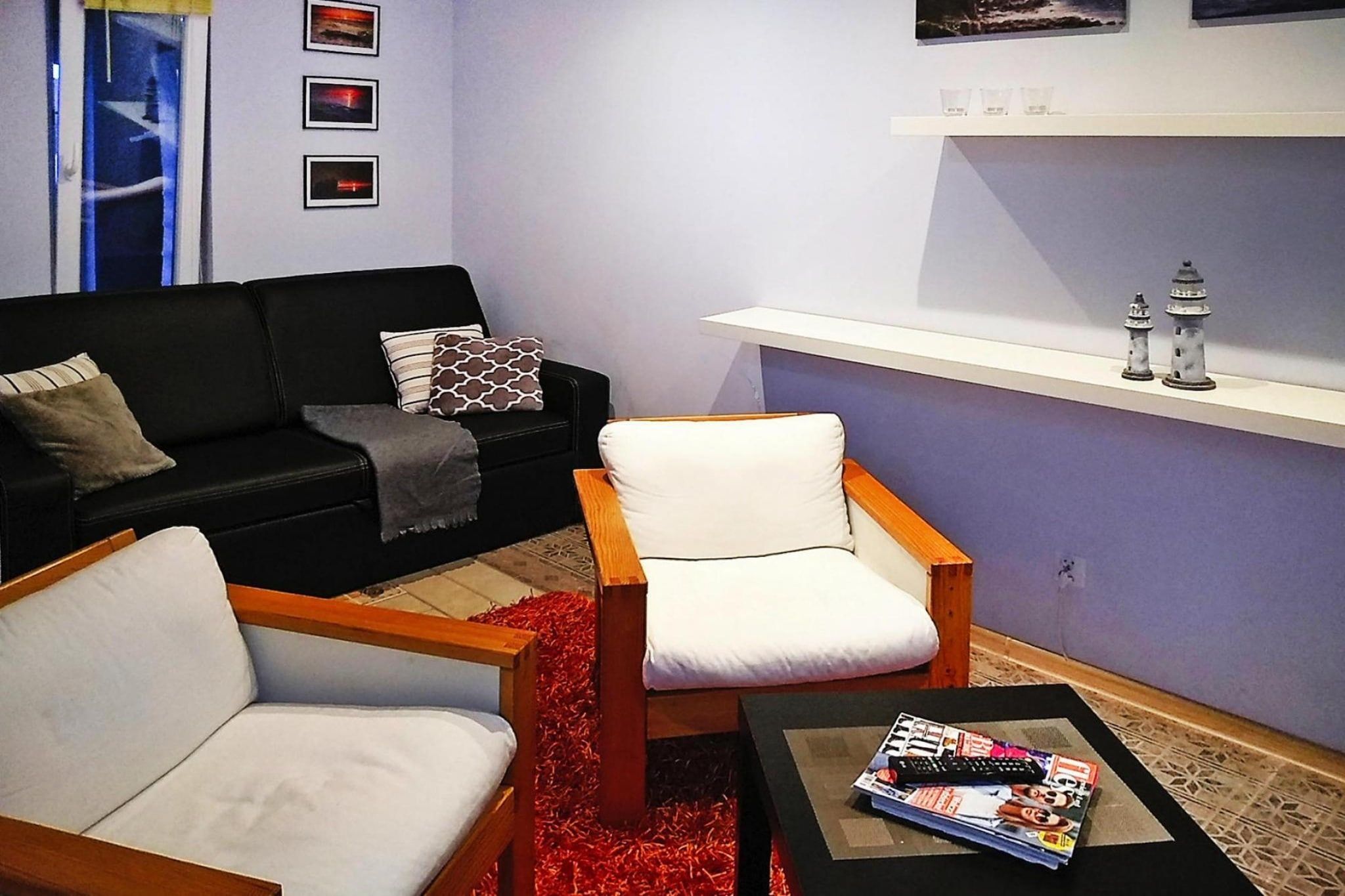 A living room with a black sofa, a white armchair, a coffee table with magazines, and a red rug.