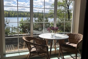 A room with a large window overlooking a lake, two wicker chairs, a small round table with a vase of flowers, and a view of trees and hills.