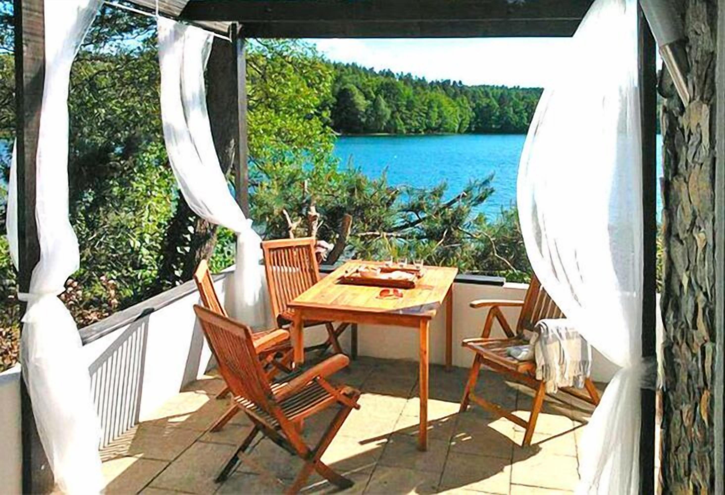 Un espacio al aire libre cubierto con mesa y sillas, con vistas a un lago y bosque.