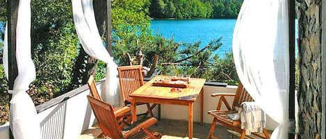 Un espacio al aire libre cubierto con mesa y sillas, con vistas a un lago y bosque.