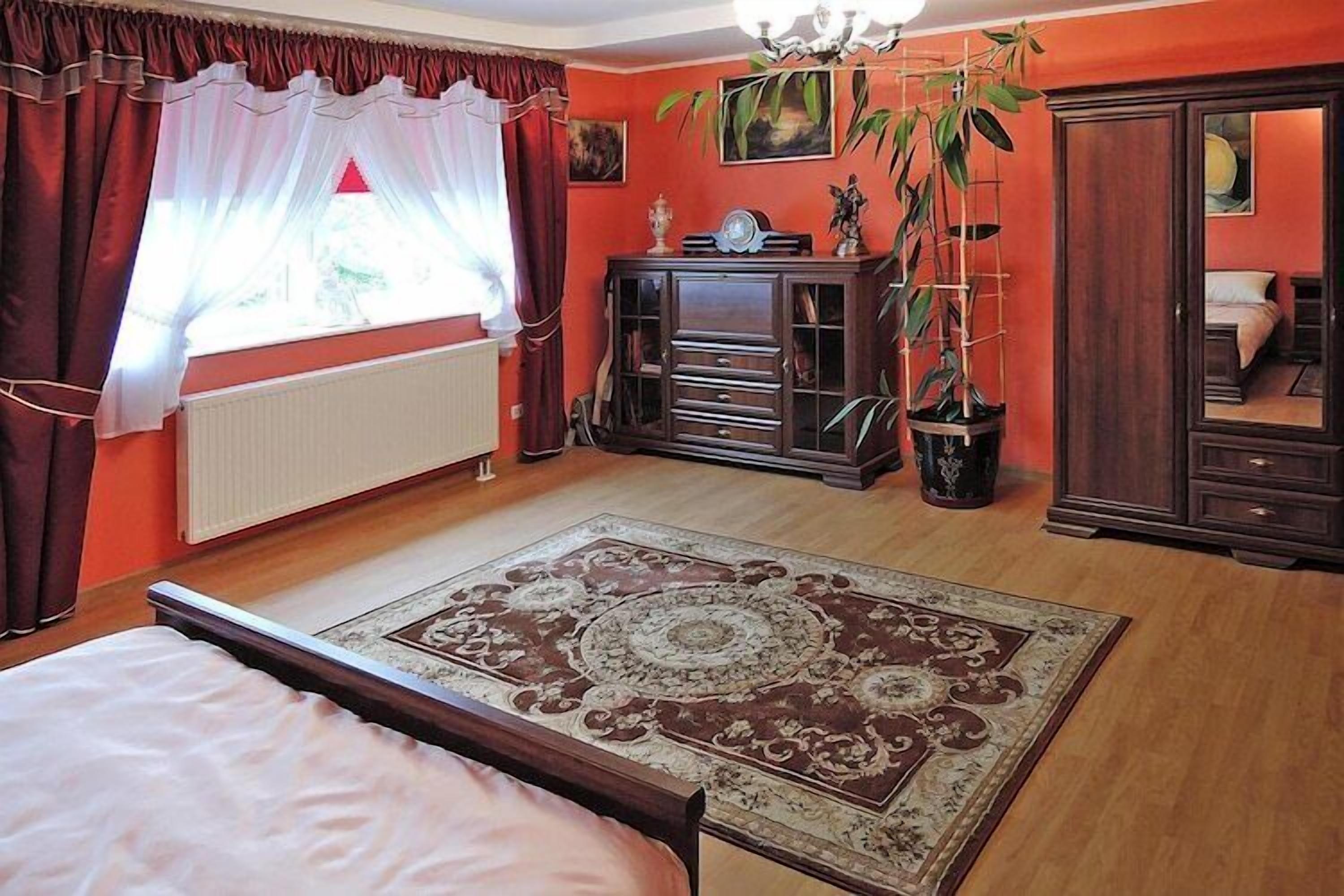 A bedroom with a bed, a wooden dresser, a radiator, a window with curtains, and a large rug.