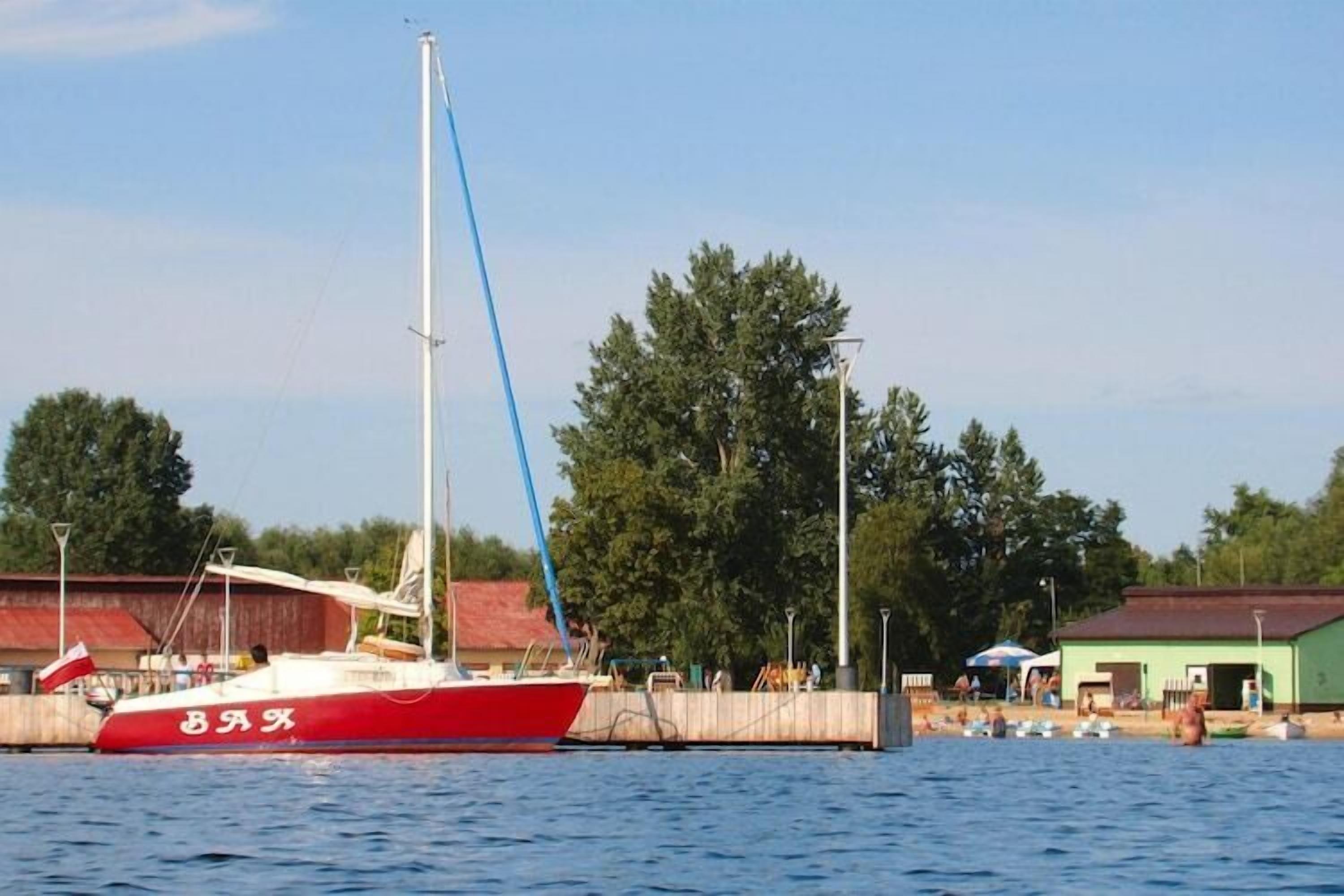 A red and white sailboat docked at a pier with a clear sky and trees in the background.