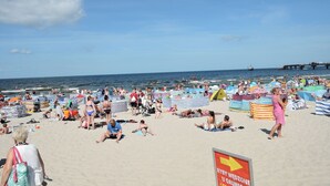 Una playa concurrida, gente tomando el sol y nadando, un muelle al fondo y un cartel de salvavidas.