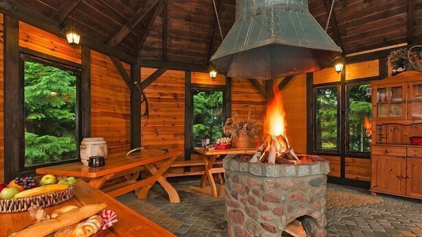 Interior rústico de una cabaña con un hogar de piedra, mobiliario de madera y grandes ventanas con vistas a la vegetación.