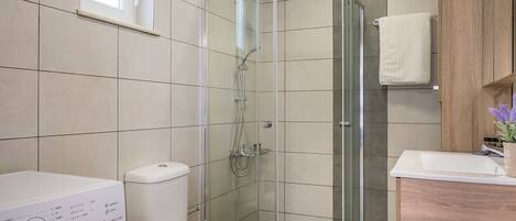 A modern bathroom with a glass-enclosed shower, a toilet, a washing machine, and a sink with a wooden cabinet.