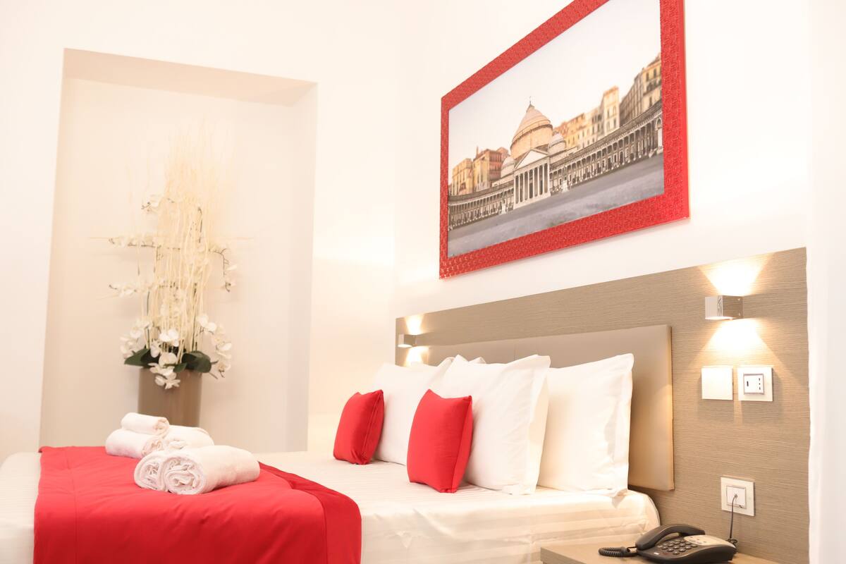 Ein Hotelzimmer mit einem Bett, rot-weiß gestreifter Bettwäsche, einer Vase mit weißen Blumen, einem gerahmten Bild an der Wand und einem Nachttischtelefon.