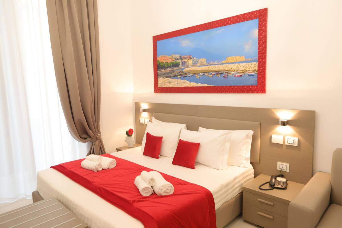 Ein Hotelzimmer mit einem Bett, roter und weißer Bettwäsche, einem Gemälde mit einer Küstenansicht, einem Nachttisch mit einem Telefon und einem Stuhl.